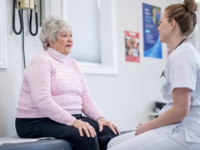 Healthcare provider in white coat sitting and talking with patient in pink sweater during medical appointment in exam room with medical equipment