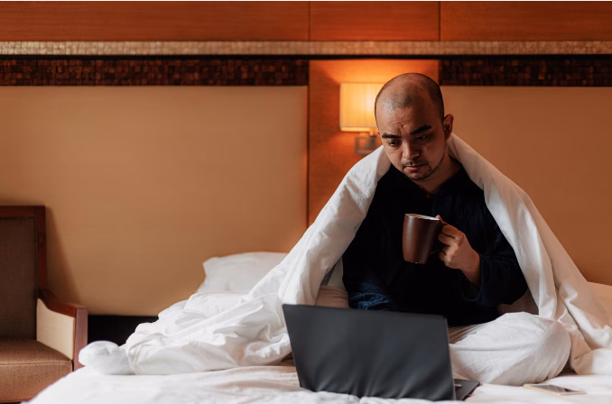 Person sitting on bed looking at laptop with concerned expression in warm-lit room