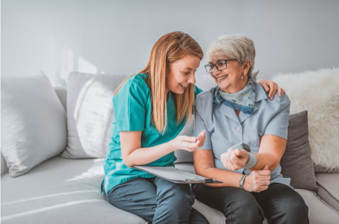 Healthcare provider in scrubs discussing blood pressure readings with an older adult patient sitting on a couch, both looking at a tablet together