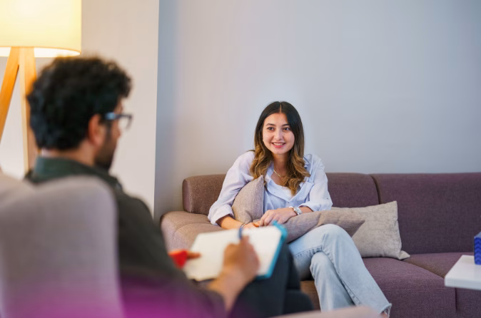Two people sit across from each other in a calm therapy office. One person sits in a chair holding a notepad while the other sits on a couch.